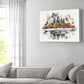 A rectangular canvas wall art print featuring a colorful watercolor-style city skyline with a reflection effect, displayed on a light gray wall above a modern light gray sofa in a living room setting.