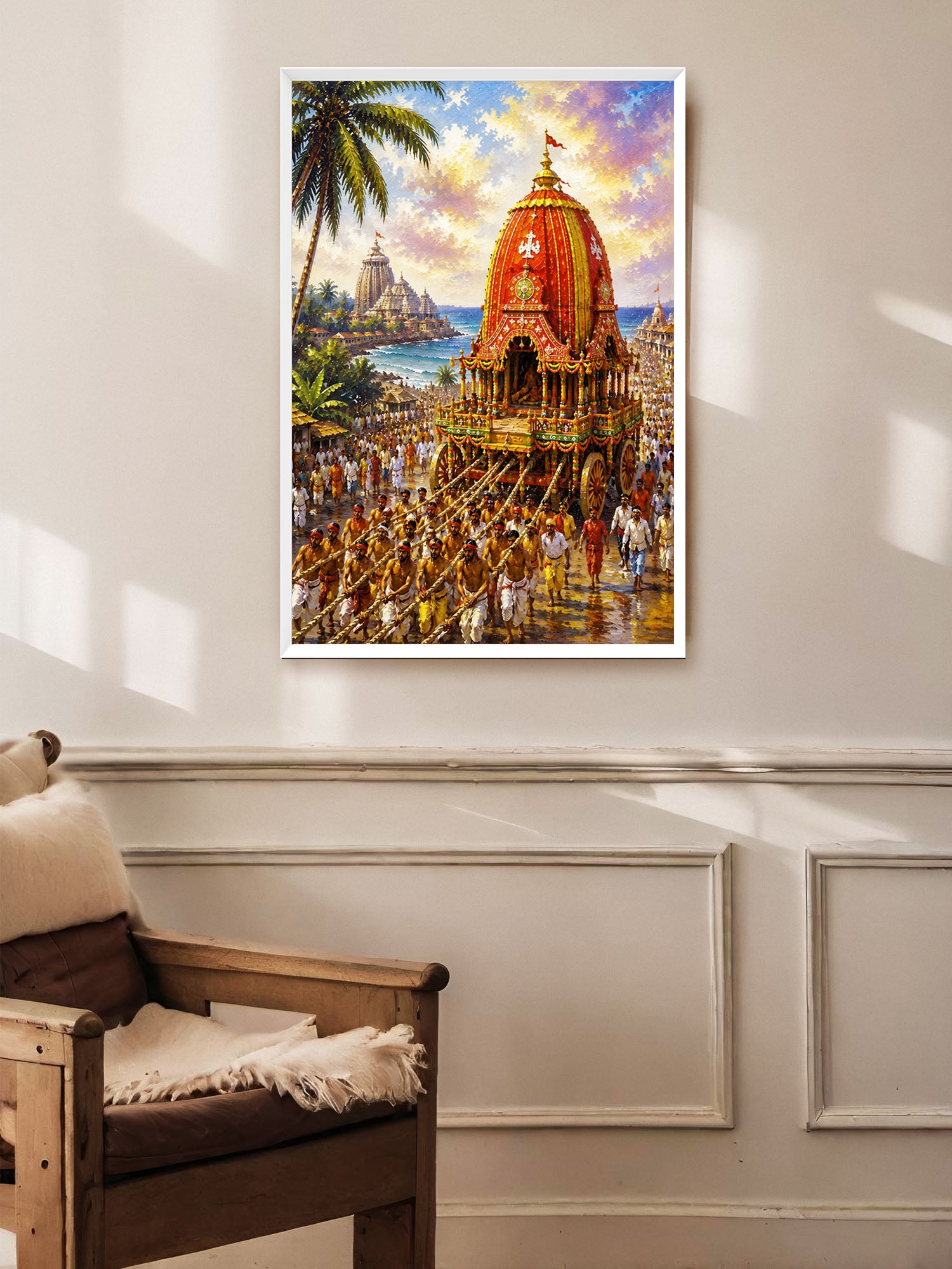 A framed colorful painting of a traditional Indian temple procession with a decorated chariot, palm tree, and sea backdrop, displayed on a beige wall above a wooden armchair.
