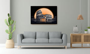 A framed wall art print depicting the Colosseum at night with a large glowing orange moon, shown hanging above a light gray sofa in a modern living room setting.