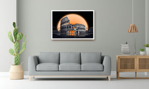 A framed wall art print depicting the Colosseum in Rome under a large orange moon, displayed on a light gray wall above a modern gray sofa in a styled living room.