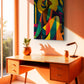 A colorful abstract canvas print of a seated figure hangs on a warm peach wall above a mid-century modern wooden desk in a sunlit room.
