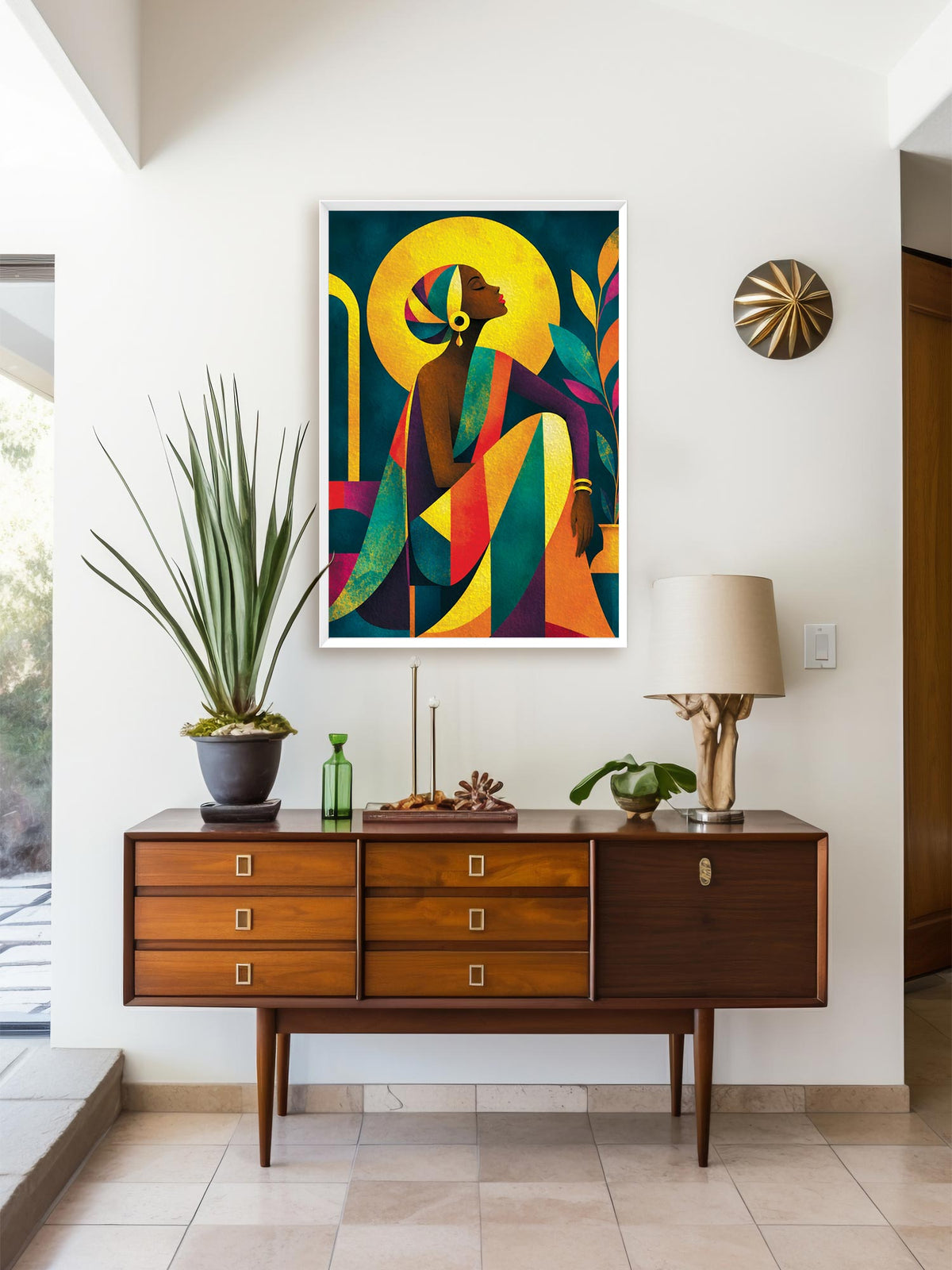 A framed colorful art print featuring an abstract figure with a yellow halo, displayed on a white wall above a mid-century modern wooden sideboard with a lamp and potted plant.