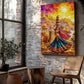 A colorful canvas wall art print depicting a vibrant folk dance scene with a woman in a blue and red skirt, camels, and a bright sunset sky, displayed on a rustic white brick wall in a styled room with a wooden chair, potted plant, and window.