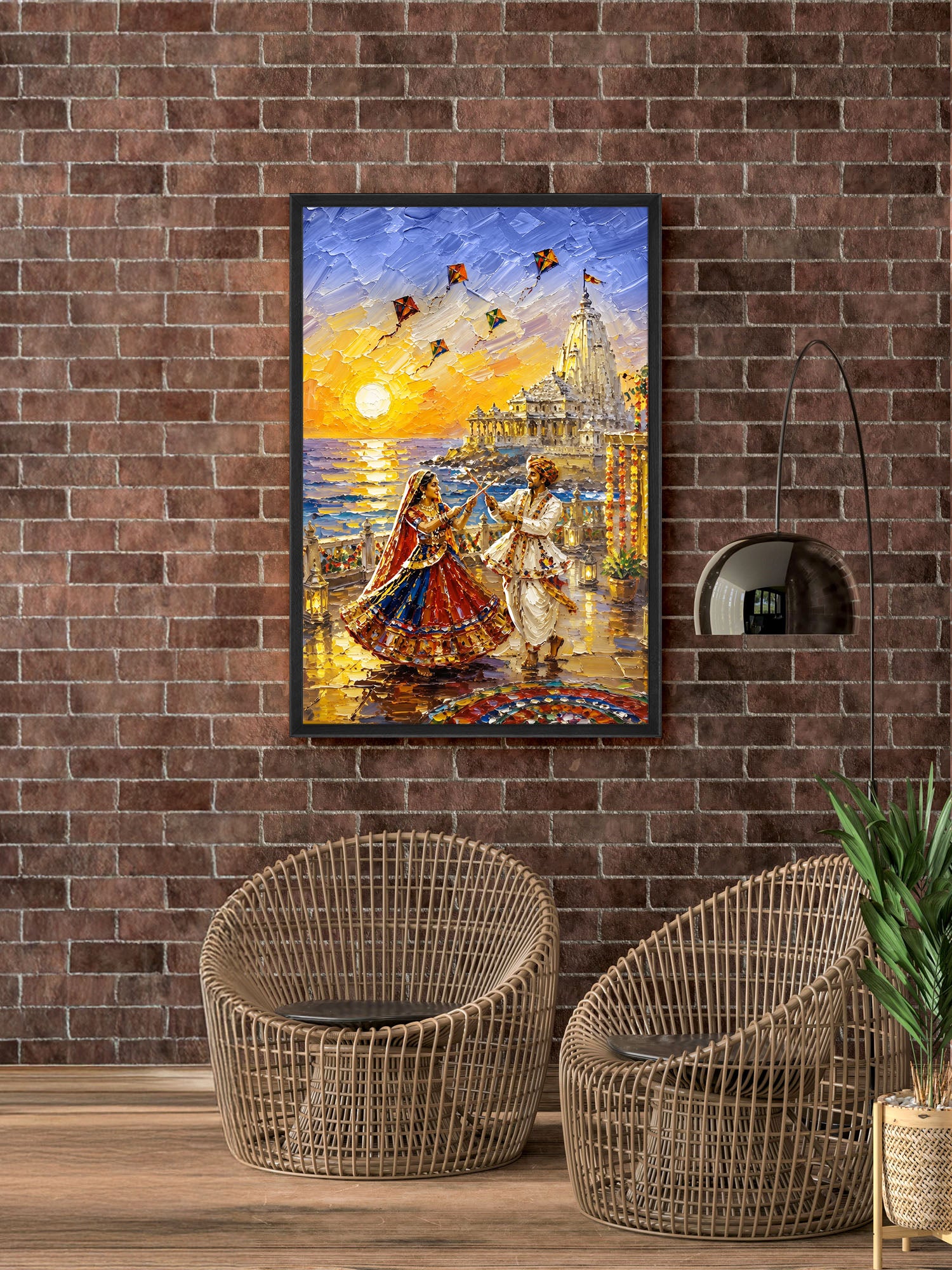 A framed colorful painting of a festive evening scene with two dancing figures, a glowing sun, and a temple-like structure, displayed on a brown brick wall above two woven armchairs.