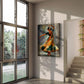 A vertical canvas art print depicting a vibrant, expressive painting of a female dancer in a flowing orange and green dress, displayed on a wall in a modern, sunlit interior room with a large window and staircase.