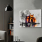 A rectangular canvas wall art print depicting a stylized illuminated monument in orange, yellow, purple, and black tones, mounted on a light gray wall in a modern living room setting with a black floor lamp, gray sofa, and shelving visible.