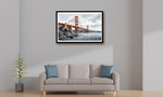 A framed wall art print of the Golden Gate Bridge hanging above a light beige sofa with blue accent pillows in a minimalist living room setting.