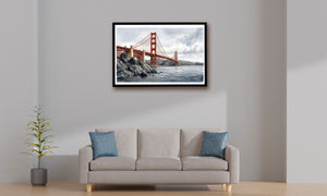 A framed wall art print of the Golden Gate Bridge hanging above a light beige sofa with blue accent pillows in a minimalist living room setting.