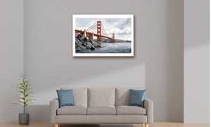 A framed art print of the Golden Gate Bridge hanging on a light gray wall above a beige sofa with blue accent pillows in a modern living room setting.