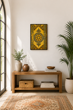 A framed square wall art print featuring a yellow and black ornate geometric medallion design, displayed in a styled room above a wooden console table with plants and books.