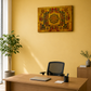 A modern home office setup featuring a light wood desk and black mesh office chair against a warm yellow wall, with a framed geometric tapestry hanging above. A potted plant stands on the left, and a small side table with a vase is on the right.