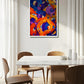 An abstract canvas painting with swirling brushstrokes of orange, purple, blue, and yellow, displayed on a light gray wall above a dining table with wooden chairs in a bright, modern room.