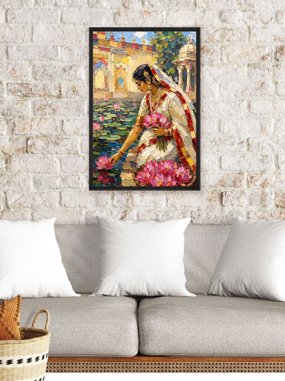 A framed vertical canvas art print depicting a woman in a white saree with pink lotus flowers, hanging on a white brick wall above a gray sofa with white pillows.