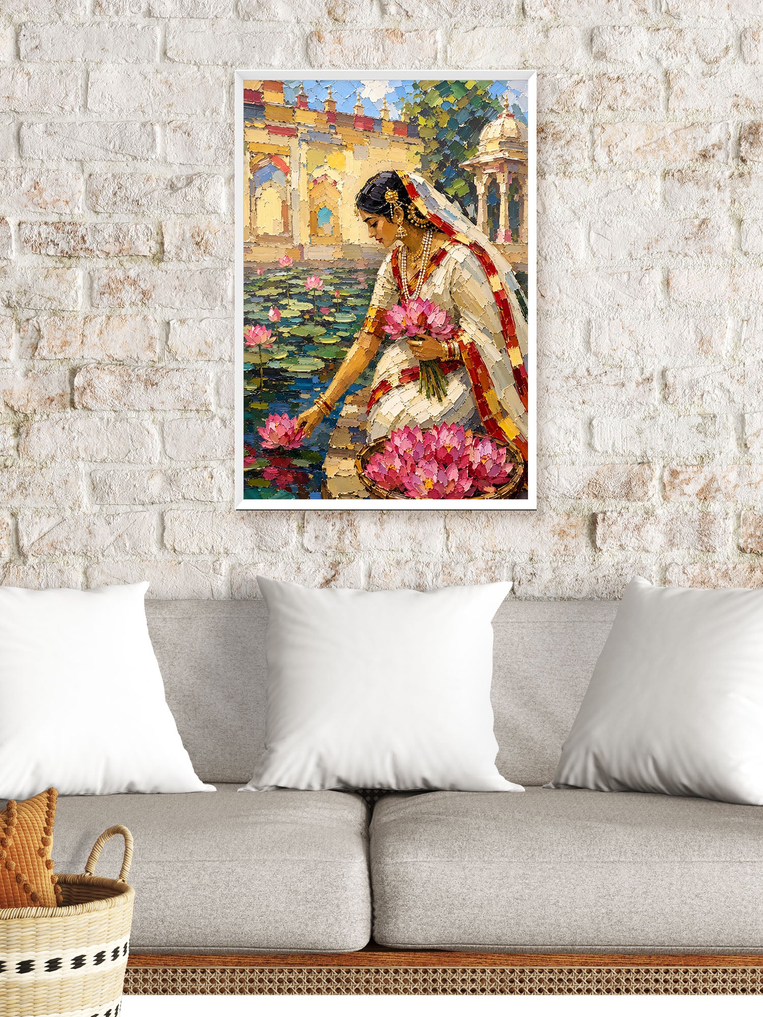 A framed vertical art print depicting a woman in a white sari with pink lotus flowers, hanging on a white brick wall above a gray sofa with white pillows.