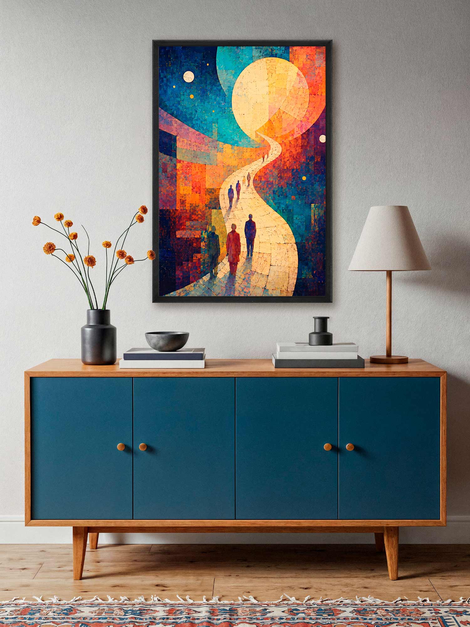 A colorful framed wall art print hangs above a mid-century modern credenza in a styled room. The artwork depicts a winding path leading to a large glowing moon, with small human silhouettes walking along it.