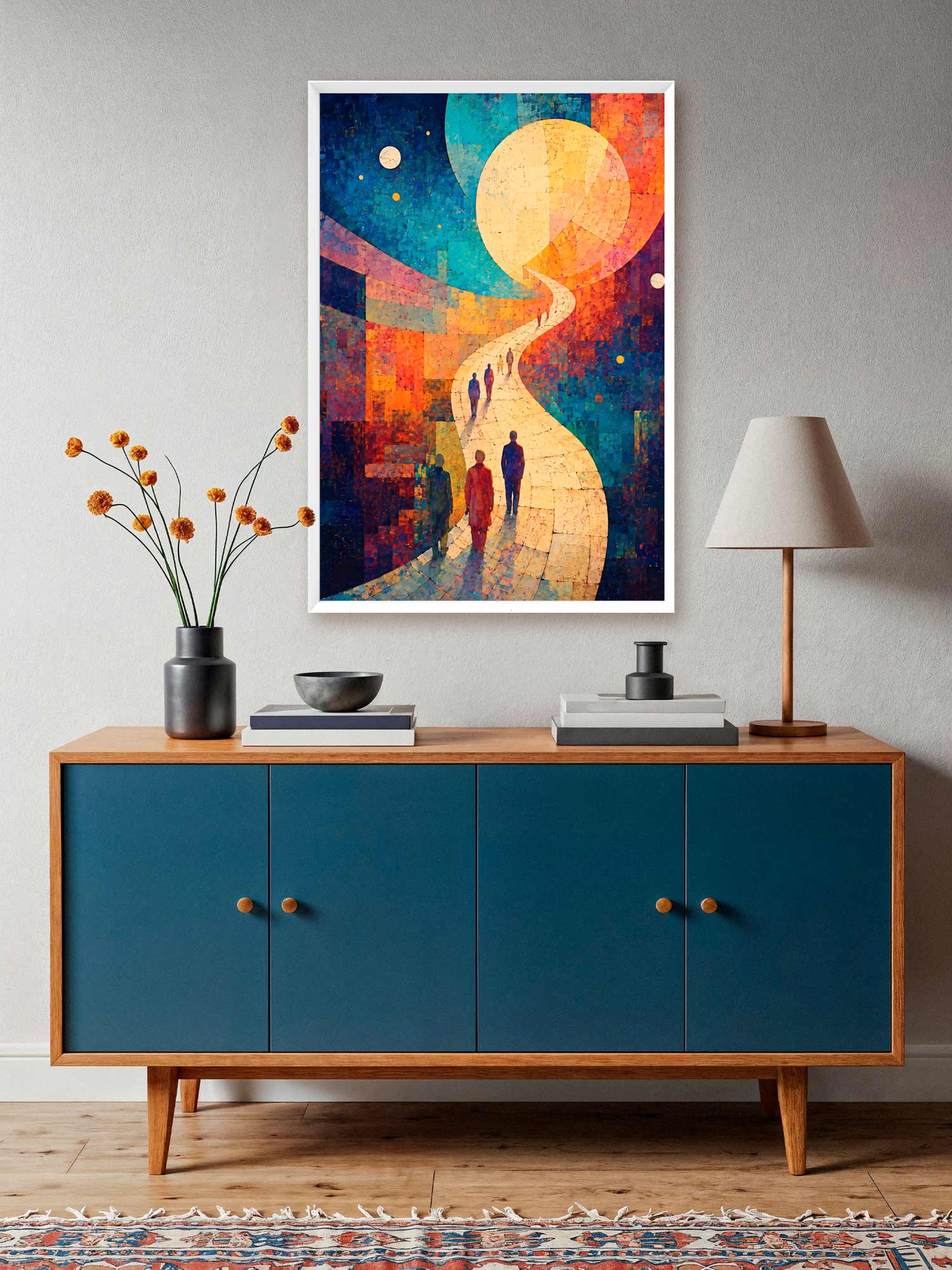 A colorful framed wall art print hangs above a mid-century modern credenza in a styled room. The artwork features an abstract scene with a large glowing moon, a winding path, and silhouetted figures, rendered in blue, orange, yellow, and red tones.