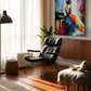 A colorful abstract canvas painting of a woman in a flowing dress hanging on a wall in a modern, sunlit living room with a black leather lounge chair and wooden floor.