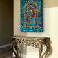 A colorful rectangular wall art print depicting a stylized archway with floral and ornamental motifs, displayed above a carved console table in a beige room.