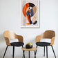 A framed abstract art print featuring orange, navy, beige, and black organic shapes, displayed on a white wall above a small wooden table with two modern wooden chairs.