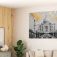 A square wall art print depicting the Taj Mahal with gold leaf accents, shown hanging on a beige wall in a modern living room setting with a sofa, wood-paneled wall, and plant.