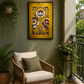A framed vertical art print featuring a golden-yellow background with purple and white floral motifs, displayed on a dark green wall in a styled patio setting with a wicker armchair, potted plants, and a small wooden table.