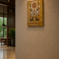 A rectangle golden canvas wall art piece with a golden-yellow border and colourful traditional folk-art style motifs, hanging on a beige textured wall in a modern interior hallway.