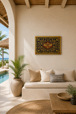 A bohemian-style framed golden wall art piece featuring a detailed central medallion and ornate borders in gold, red, and blue, hanging on a cream stucco wall above a white cushioned daybed with neutral pillows in a sunlit patio setting.