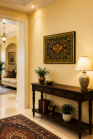A framed ornate oriental-style wall hanging with a central medallion, displayed in a warm, elegant living room above a dark wood console table with a lamp and plants.