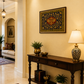 A warm, elegant interior hallway featuring a dark wood console table decorated with a patterned table lamp, potted plants, and a decorative rug. On the cream-colored wall above the console, a framed oriental-style tapestry hangs.