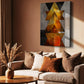 A vertical abstract canvas print with geometric shapes in gold, orange, black, and gray, displayed on a warm brown wall above a beige sofa with decorative pillows in a styled living room.
