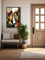 A framed abstract art print featuring two stylized faces hangs on a beige wall in a modern entryway. The scene includes a light wood bench with pillows, a potted green plant, a wood door with glass panes, and a light rug on a wood floor.