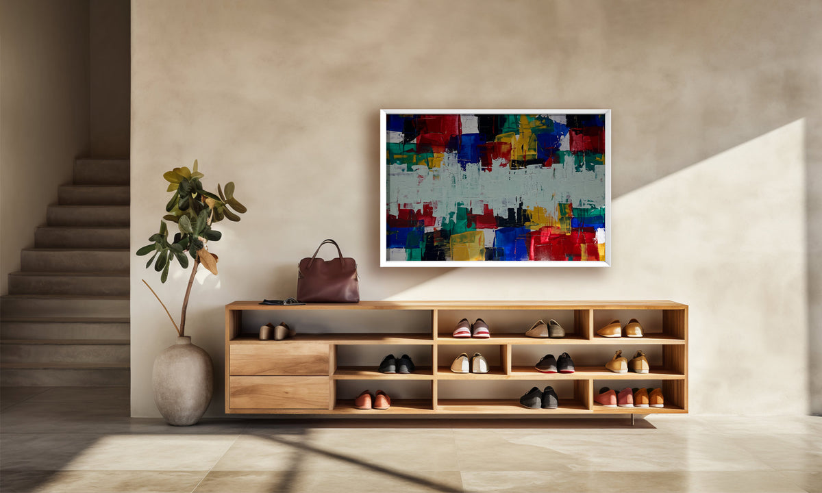 A colorful abstract canvas print with a white frame hangs on a beige wall above a wooden shoe cabinet in a modern, sunlit entryway.